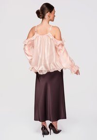 Femme aux cheveux relevés en chignon, portant un chemisier en satin rose pâle, épaules dénudées, et une longue jupe en satin marron foncé avec des talons noirs.