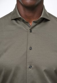 Camicia verde oliva con bottoni, realizzata in tessuto a trama. Caratterizzata da un colletto a punta e due bottoni neri sul davanti. Tessuto liscio e leggero.