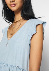 Vaaleansininen chambray-mekko, jossa on V-kaula-aukko, röyhelöhihoja ja kerroksellinen muotoilu. Kultaketjuiset kaulaketjut ja riipus lisäävät yksityiskohtia.