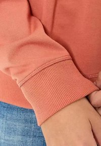 Sweatshirt corail à manches longues avec un poignet côtelé. Le tissu semble doux et lisse, présentant des coutures détaillées le long des bords.