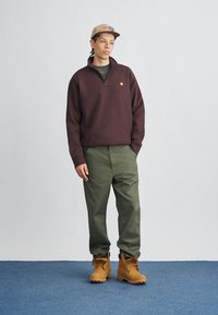 Jeune personne portant une casquette couleur fauve, un pull à demi-fermeture éclair marron foncé, un pantalon vert olive et des bottes de travail couleur fauve, debout sur un tapis bleu devant un mur gris.