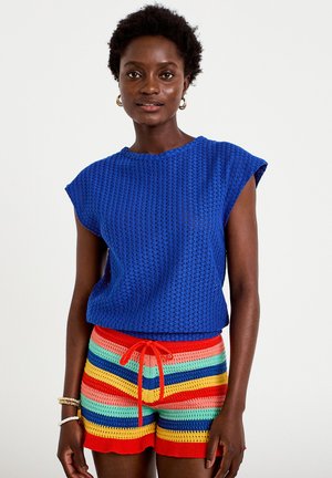 Jonge vrouw draagt een blauwe mouwloze gebreide top en multicolor gestreepte gebreide shorts met een rode trekkoord, staand tegen een effen achtergrond.