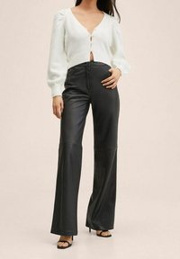 Bílý pletený cardigan s nafouknutými rukávy a knoflíkovými detaily, v kombinaci s černými vysokými kalhotami z umělé kůže s rozšířenými nohavicemi a černými sandály na podpatku.