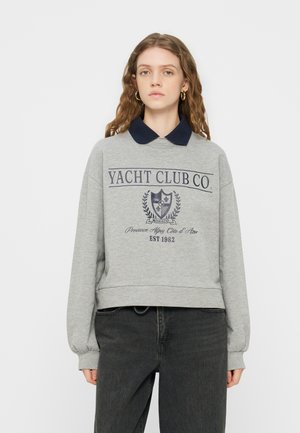 Ung kvinde med bølget hår iført grå sweatshirt med mørkeblå krave og "Yacht Club Co."-tryk, kombineret med sorte jeans, stående mod en hvid baggrund.