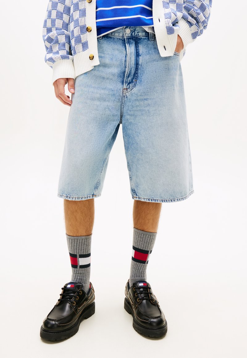 Άνδρας που φοράει ανοιχτό μπλε τζιν σόρτς μέχρι το γόνατο, γκρι κάλτσες με σχέδια και μαύρα δερμάτινα παπούτσια με κορδόνια.