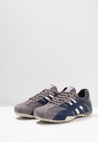 Chaussures de sport grises et bleu marine avec un mélange de matériaux en daim et en mesh. Elles présentent des bandes blanches, une semelle extérieure en caoutchouc et un laçage à l'avant.