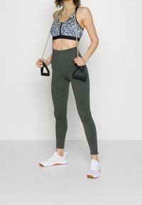 Sportowy stanik w czarno-białym wzorze kwiatowym; wysokie oliwkowe legginsy; białe trampki z pomarańczowymi akcentami; trzyma taśmy oporowe.