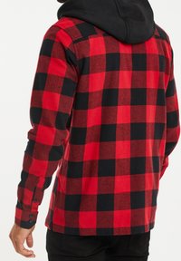 Man in een rood en zwart geblokt flanellen shirt met een zwarte capuchon, van achteren weergegeven, tegen een effen witte achtergrond.
