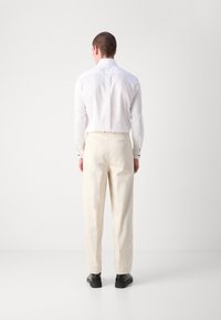 Camisa blanca de manga larga con cuello clásico, pantalones de color crema y zapatos negros, vistos desde atrás, contra un fondo liso.
