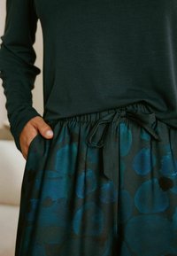 Dunkelgrünes langärmliges Oberteil, kombiniert mit einem gemusterten Rock mit blauen und türkisfarbenen Kreisdesigns, elastischem Bund und einem Bindedetail.