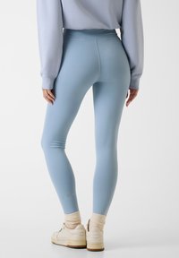 Leggings bleu clair en matière extensible, avec une taille haute et un petit logo. Portés avec des baskets blanches et des chaussettes beige.