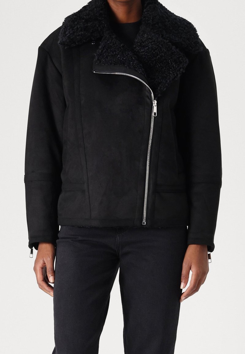 Personne portant une veste en shearling noire avec un grand col et une fermeture éclair asymétrique argentée, associée à un pantalon noir.