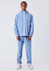Lichtblauwe trainingspak van soepel, lichtgewicht materiaal. Heeft een capuchon, witte ritssluiting en contrasterende witte stiksels op zowel de jas als de broek.