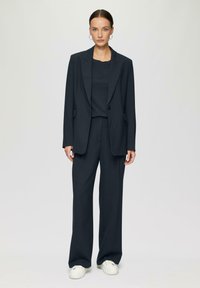 Dunkelgrauer, taillierter Blazer mit Reverskragen, passenden weit geschnittenen Hosen und einem strukturierten Rundhalsshirt, kombiniert mit weißen Sneakers.