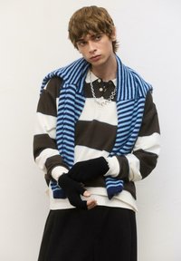 Maglione a righe blu e nere appoggiato su un polo scuro con righe bianche e marroni. Guanti neri senza dita e una collana a catena completano il look.