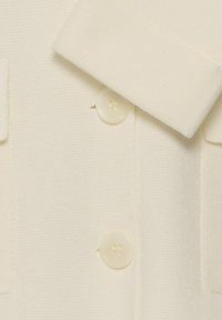 Gros plan sur un tissu texturé crème avec deux grands boutons crème et un revers ou un détail de col plié.