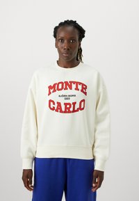 Tejfehér pulóver piros "MONTE CARLO" felirattal és logóval, kerek nyakkivágással és bordázott mandzsettával, kék nadrággal párosítva.