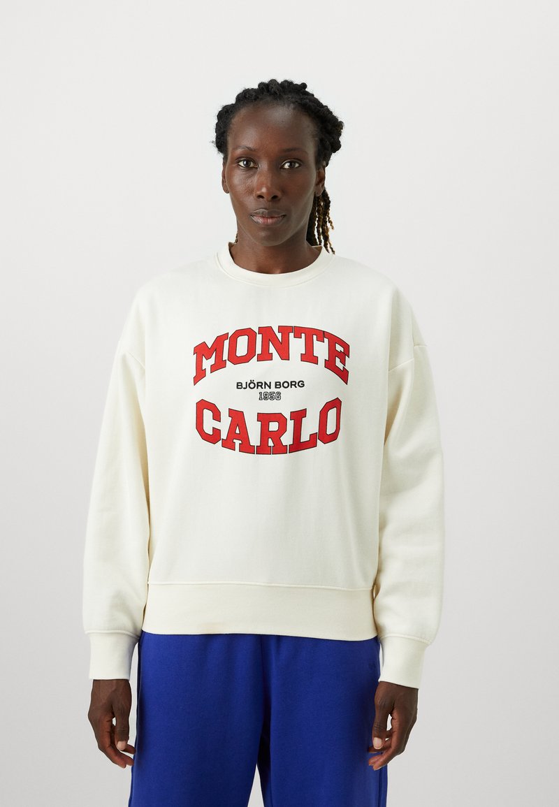 Tejfehér pulóver piros "MONTE CARLO" felirattal és logóval, kerek nyakkivágással és bordázott mandzsettával, kék nadrággal párosítva.