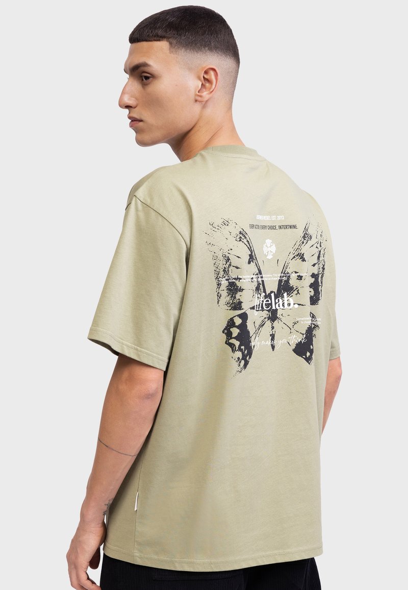 T-shirt en coton vert olive avec un grand papillon noir imprimé dans le dos, accompagné de texte stylisé au-dessus et en dessous du motif.