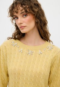 Maglione giallo lavorato a maglia con motivo a treccia, decorato con disegni floreali impreziositi da gemme trasparenti e perle finte attorno al collo.