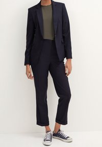 Marineblazer und Hosenanzug mit einem taillierten olivfarbenen Oberteil, das umgeschlagene Bündchen aufweist, sowie lässigen marineblauen Sneakers, die einen eleganten, aber entspannten Look kreieren.