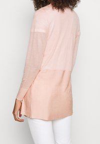 Dlouhý rukáv, lehký pletený svetr v jemně růžové barvě s žebrovanou texturou na dolní polovině a hladkým povrchem na horní části.