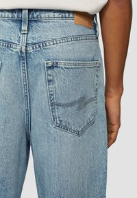 Jean en denim bleu clair avec une poche arrière ornée de coutures décoratives et un patch en cuir marron sur la ceinture.