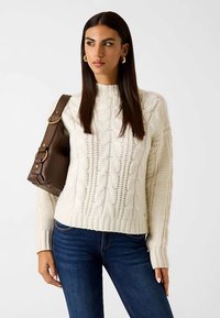 Pull crème en maille torsadée avec ourlet côtelé et encolure ras-du-cou. Porté avec un jean denim foncé, tenant un sac à bandoulière marron.