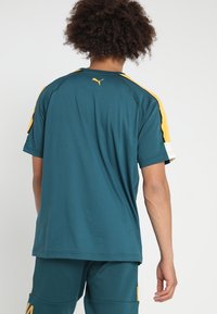 Turkos tränings t-shirt med gula detaljer på axlarna, tillverkad av lättviktigt material, har en rundad halsringning och en liten logotyp på baksidan.