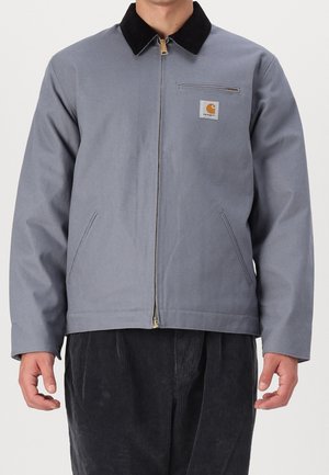 Man wearing light gray zip-up jacket with black collar, chest zipper pocket, and Carhartt logo patch, paired with black corduroy pants.