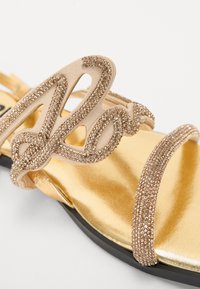 Gold flat sandal featuring a suede strap with decorative rhinestones and a metallic gold footbed. Black outsole and minimalistic design.