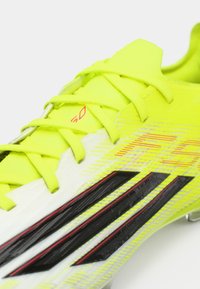 Φωτεινό κίτρινο ποδοσφαιρικό παπούτσι με υφασμένη συνθετική επάνω επιφάνεια, μαύρες και κόκκινες λεπτομέρειες και κλείσιμο με κορδόνια. Διαθέτει χαρακτηριστικό ριγέ σχέδιο.