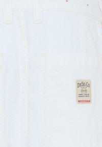 Białe ogrodniczki z denimu z małą, beżową metką marki z niebieskim napisem na kieszeni. Projekt zawiera widoczne przeszycia i kieszenie.