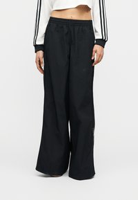 Oseba, oblečena v črne široke športne hlače in bel top z dolgimi rokavi, prirezanimi na trebuh, s črnimi črtami na rokavih, stoji pred belim ozadjem.