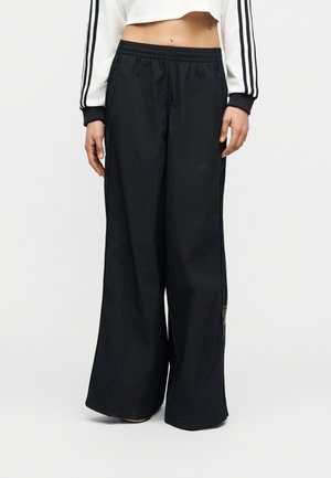 Persona, kas valkā melnas plašas sporta bikses un baltu īsu garām piedurknēm augšu ar melnām svītrām piedurknēs, stāvot pret balto fonu.