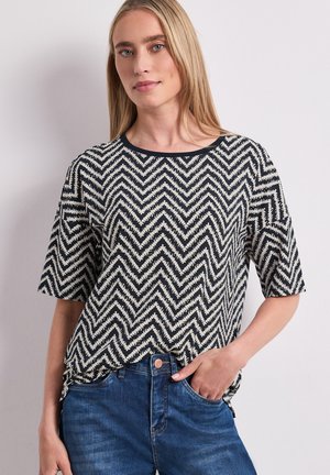 Femme aux cheveux blonds raides portant une chemise à manches courtes noire et blanche à motif zigzag et un jean bleu, main dans la poche.