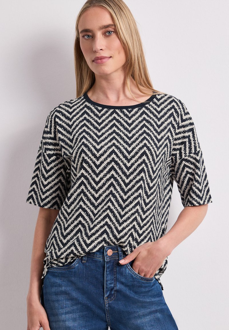 Femme aux cheveux blonds raides portant une chemise à manches courtes noire et blanche à motif zigzag et un jean bleu, main dans la poche.