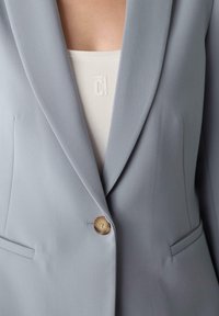 Blazer grigio chiaro con una texture liscia, un singolo bottone dorato e ampie rever. Il top interno bianco mostra un logo sottile.