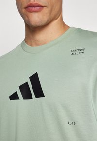 Světle zelené bavlněné tričko s černými diagonálními pruhy a textem "TRAINING ALL_GYM" na hrudi, kulatý výstřih a měkká textura.