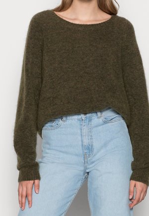 Femme portant un pull vert foncé ample et duveteux rentré dans un jean taille haute bleu clair, vue du cou aux hanches.