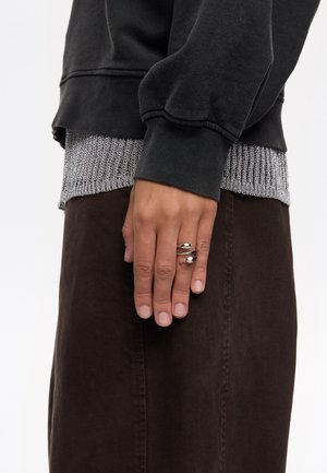 Sudadera negra con dobladillo de ribete plateado, superpuesta a una falda marrón. Mano con anillos de plata descansando sobre la tela de la falda.