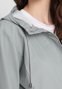 Jasnoszara wodoodporna kurtka z kapturem, wyposażona w ściągacz, gładką teksturę oraz srebrne elementy ozdobne.