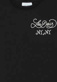 Black cotton t-shirt featuring white cursive text "Les Deux" and "NY, NY" on the upper left side. Smooth texture, round neckline.