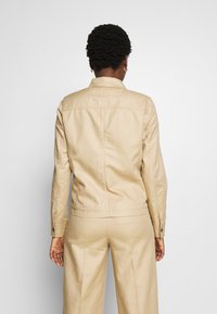 Giacca leggera beige con colletto, maniche lunghe e dettagli con bottoni. Tessuto dalla superficie liscia, design aderente e stile minimalista.