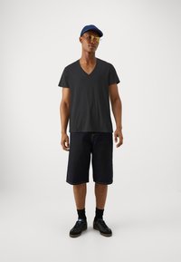 Tricou negru cu decolteu în V, pantaloni scurți din denim închiși la culoare, până la genunchi, cu cusături vizibile, adidași negri și o șapcă albastră cu ochelari de soare.