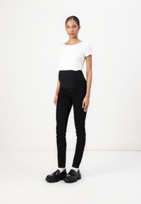 Camiseta blanca cortada, pantalones negros cómodos para embarazadas con cintura elástica, y mocasines negros gruesos, sobre un fondo liso.