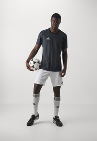 Mørkegrå trenings T-skjorte med korte ermer, med en liten hvit logo, parret med hvite shorts og kne-høye sokker med svarte striper. En fotball holdes i den ene hånden.