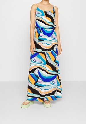 Vrouw draagt een mouwloze, enkelhoge jurk met een kleurrijk abstract patroon in blauw, oranje, zwart en wit, gecombineerd met lichtgroene sandalen met plateauzool.