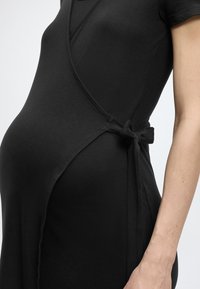 Κοντινό πλάνο ενός εγκύου ατόμου που φοράει εφαρμοστό μαύρο φόρεμα τύπου wrap δεμένο στη μέση με φιόγκο.