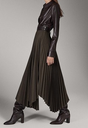 Femme portant une chemise en cuir marron foncé, une jupe plissée asymétrique taille haute et des bottes marron à talons mi-mollet.